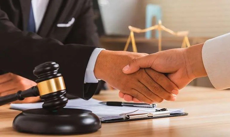 Apretón de manos sobre un escritorio con un contrato, un martillo de juez y una balanza.