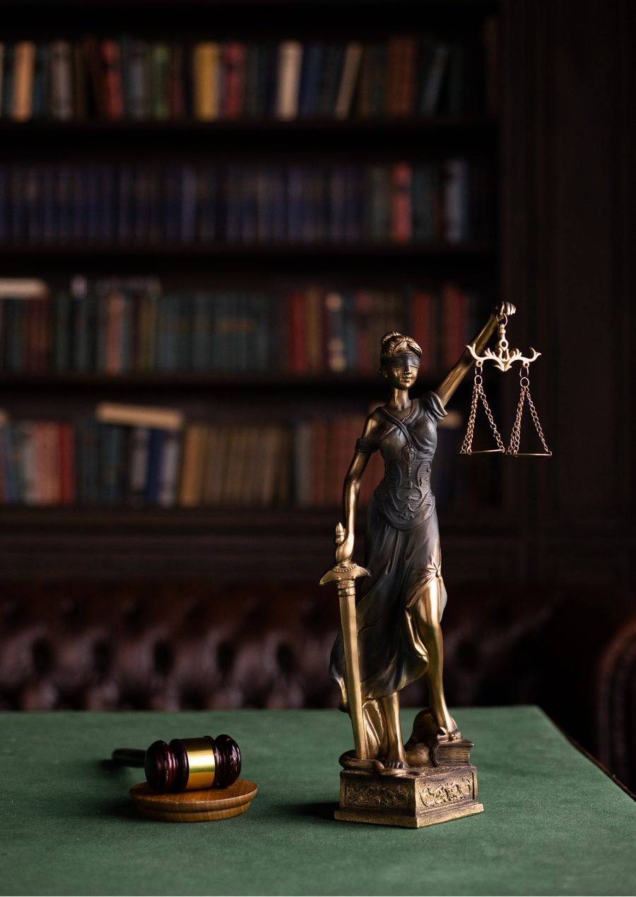 Estatua de la Justicia con balanza y espada sobre un escritorio, frente a una biblioteca.